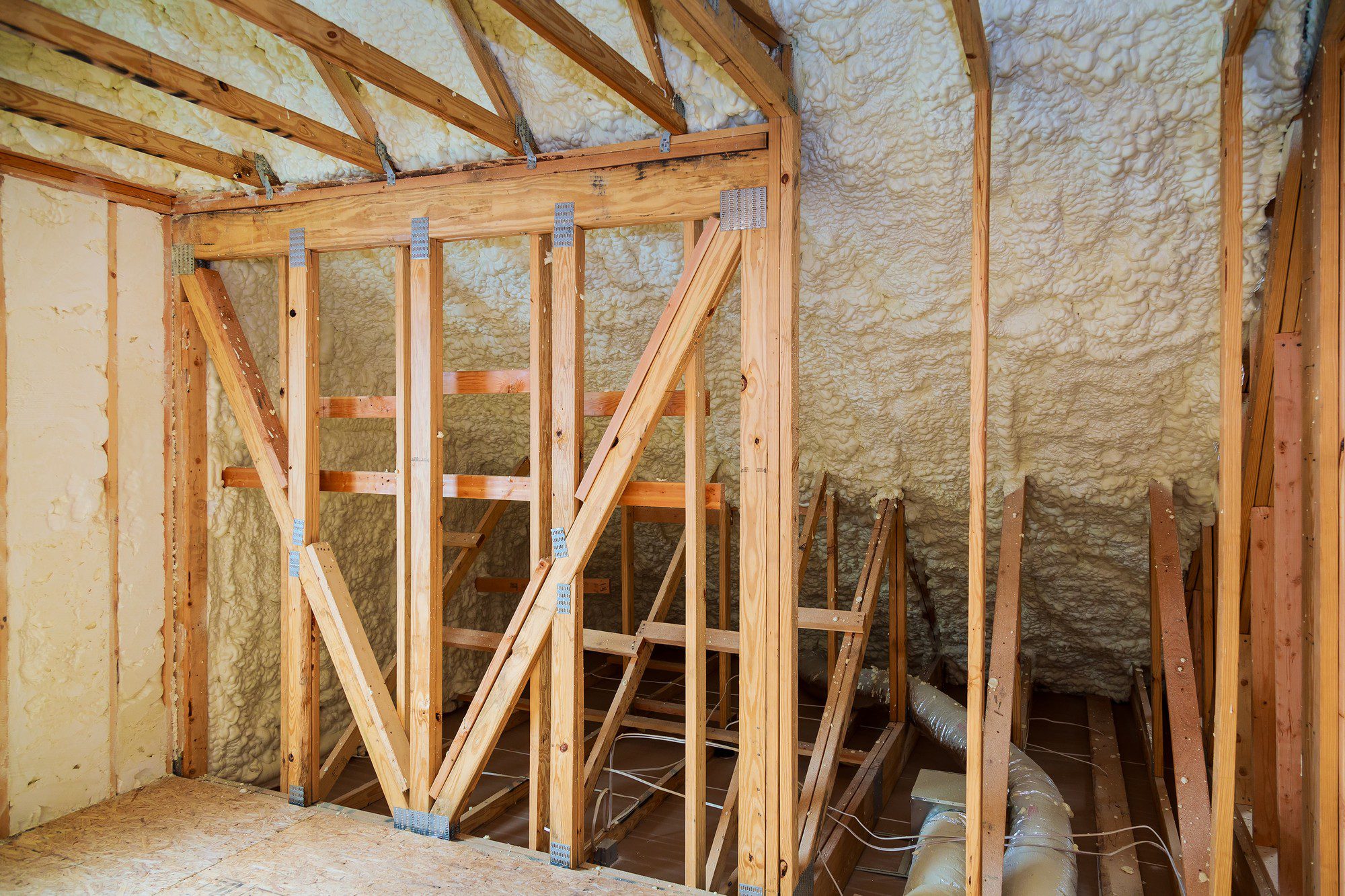 This image shows the interior framework of a building under construction or renovation. There are wooden studs, joists, and rafters making up the structural framework. Spray foam insulation has been applied to the walls and ceiling for thermal insulation, as evident by the expanding foam texture that covers these surfaces. The wooden flooring provides a base for walking, and there appears to be some ductwork, possibly for HVAC (Heating, Ventilation, and Air Conditioning) systems, running along the floor between the joists. Additionally, there are some metal ties and strapping used to reinforce the wooden structure. Overall, this appears to be an attic or upper-level space in the process of being finished or remodeled.