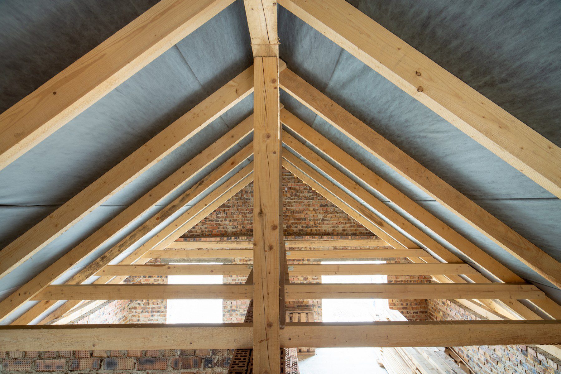 This image shows the interior framework of a pitched roof of a building, seen from the inside. The structure is composed of wooden beams or rafters that come together in a triangular shape, which is typical for providing support under the roof covering. The framework is set against a backdrop of an exposed brick wall, and it looks like there might be insulation material installed between the rafters, as indicated by the gray layers seen in between the wood. The image also shows a clear view of the wooden ridge beam at the peak, where the rafters meet. The combination of wooden structure and brick wall gives the impression of a loft or an unfinished attic space in a building that may be undergoing renovation or construction.
